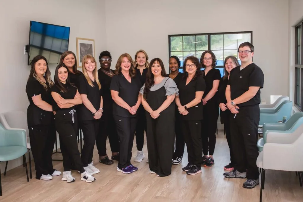 Twelve individuals pose in a modern clinical office; eleven wear black scrubs and one wears a gray top with black pants, suggesting mixed roles; background includes light wood flooring, pastel chairs, large windows, and wall-mounted TV.