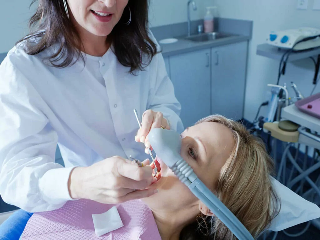 A dental professional performs a procedure on a patient in a dental chair, wearing a pink dental bib and a nasal mask, surrounded by dental office equipment.