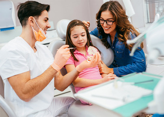 A dentist in a white uniform and orange gloves holds a dental model, explaining its features to a child seated in a dental chair, accompanied by an adult providing support.