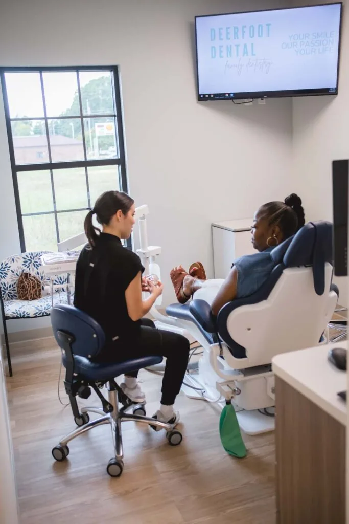 A dental professional interacting with a patient in a modern, well-lit dental office with wooden flooring and a large window in the background.