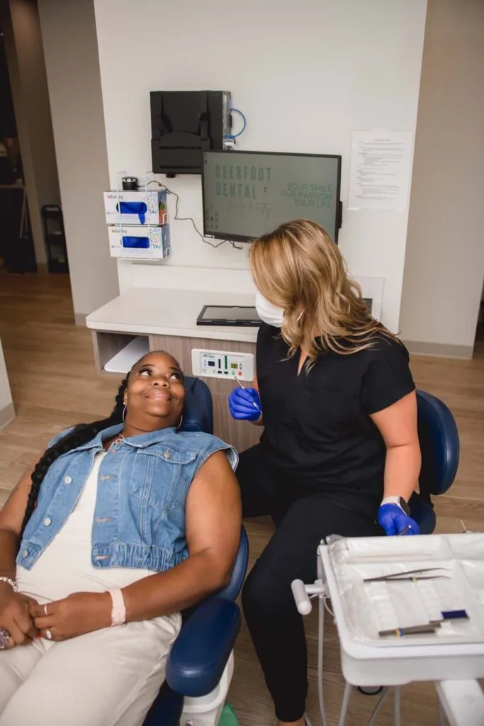 A dental professional in black scrubs and blue gloves attends to a patient seated in a dental chair, with a tray of instruments nearby. The background features a monitor displaying "DEERFOOT DENTAL Your Smile Is Our Top Priority," wooden flooring, and dental equipment mounted on the wall.