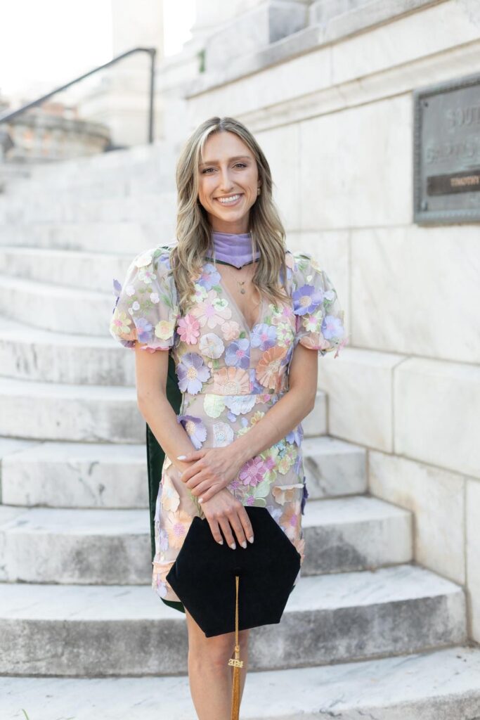 Dr. Bidikov standing confidently on marble stairs, wearing a pastel floral puff-sleeve dress and holding a black graduation cap with a gold tassel. A stone wall and commemorative plaque form the background, highlighting an academic milestone.